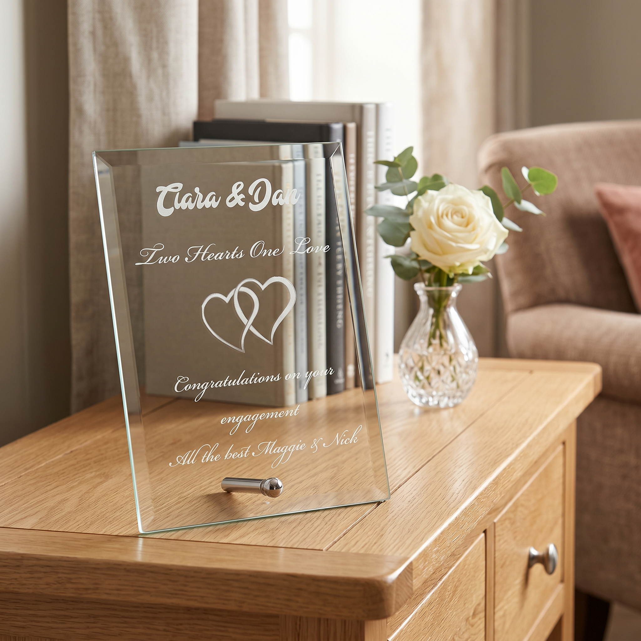 A glass frame with a message that reads "Clara & Dan" and "Two Hearts One Love" is placed on a wooden table. Next to the frame is a vase with a single white rose, and there are several books and a lamp in the background.