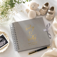a grey baby book with gold text, a stuffed teddy bear, a vase with flowers, baby shoes, and a pen on a white surface.