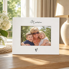 A white picture frame with a photo of a woman and two children inside, placed on a wooden surface near a window.