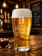 A glass of beer with the number 18 and the text "BRANDON HAPPY BIRTHDAY" printed on it, placed on a wooden table in a bar setting.