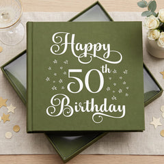 A green photo album with the text "Happy 50th Birthday" printed on it, placed on a table with a glass of wine and a bouquet of flowers.