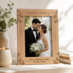 A wooden picture frame with a photo of a couple, presumably a bride and groom, inside it. The frame has a floral design around the edges and the couple is smiling at each other. The frame is placed on a table next to a vase with flowers and a lit candle.