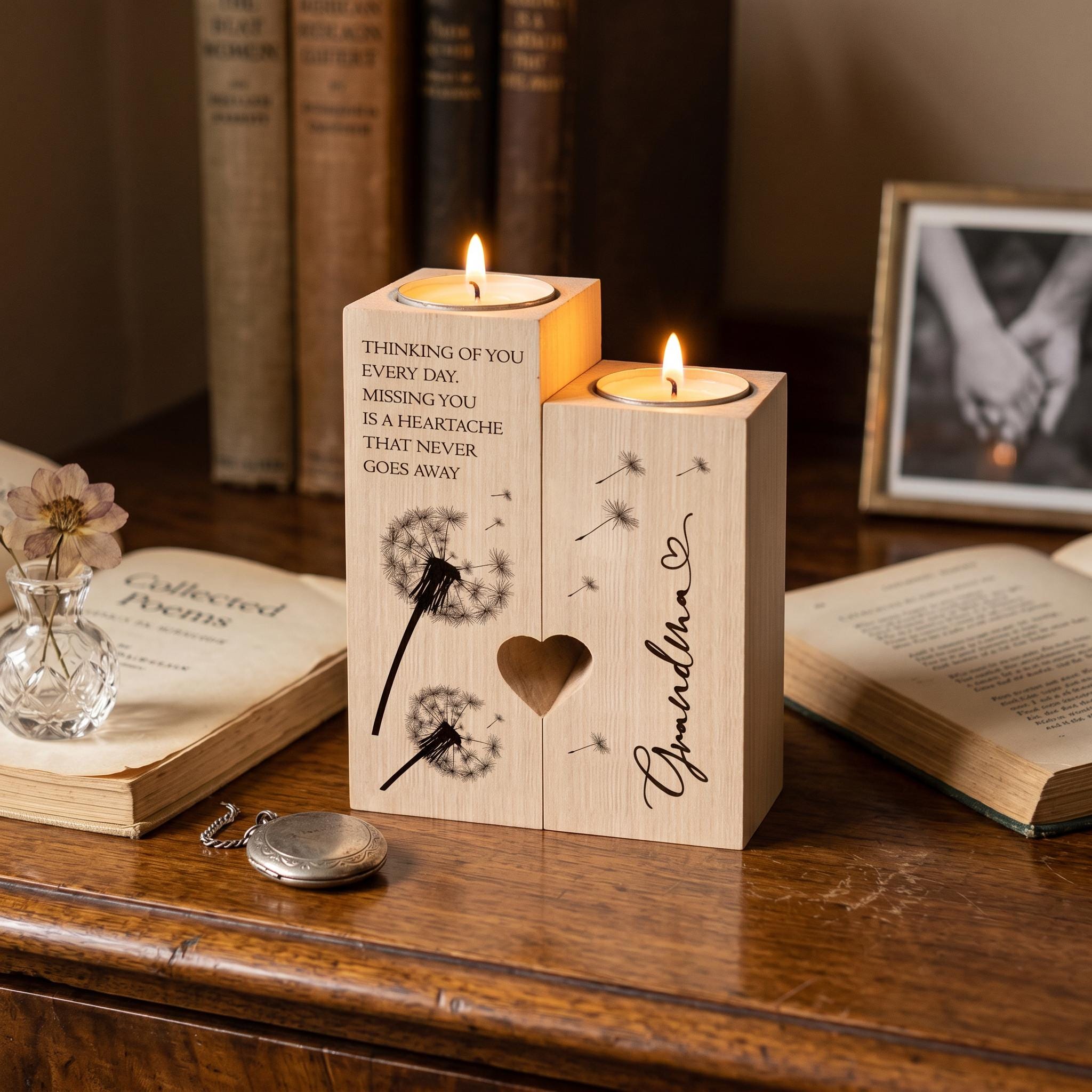 A wooden candle holder with two lit candles, featuring a heart-shaped cutout and dandelion designs, sits on a wooden surface. The candle holder also has a message that reads "Thinking of you every day, you are a heartache that never goes away". In the background, there are books and a framed photograph, creating a cozy and thoughtful atmosphere.