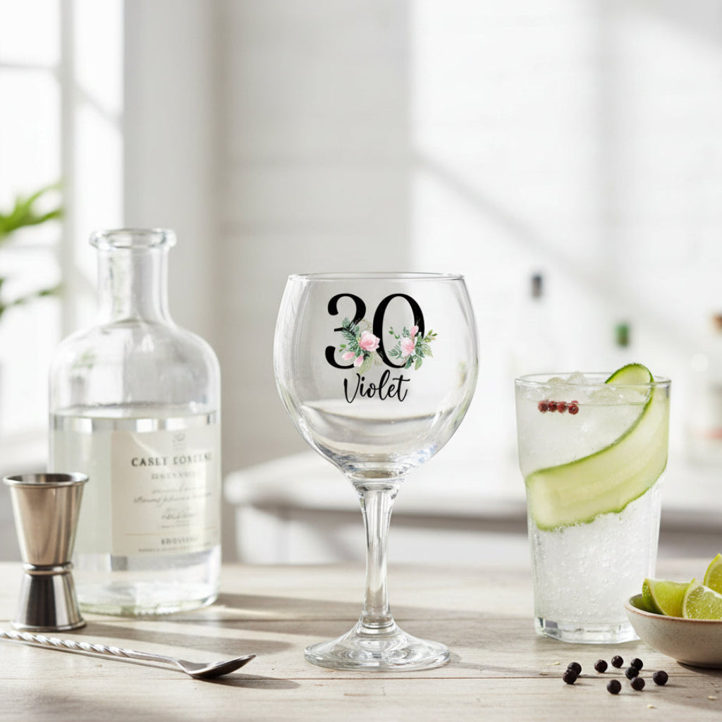 30th Birthday Gin glass with floral design and text on a table with a bottle and glass of water.
