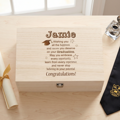 A wooden box with a graduation cap and mortarboard on it, along with a graduation gown and a graduation cap, sits on a wooden surface.