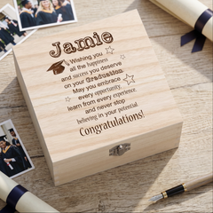 A wooden box with a personalized message engraved on it, placed on a wooden surface alongside graduation caps and gowns.