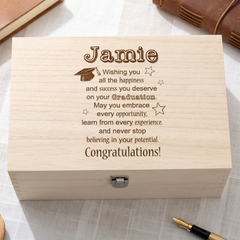 A wooden box with a personalized message engraved on it, placed on a surface with a pen and a book in the background.