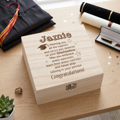 A wooden box with a personalized message engraved on it, placed on a wooden surface alongside a graduation cap, a graduation gown, and some confetti.