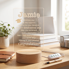 a wooden desk with a wooden stand displaying a personalized graduation message, a pen, a book, and a plant.