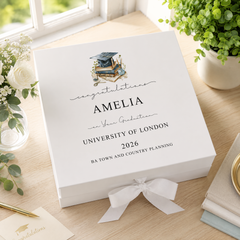 a white graduation box with a graduation cap and mortarboard, placed on a wooden surface next to a potted plant and some books.