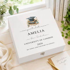 a white graduation box with a graduation cap and mortarboard, placed on a wooden surface next to some books and a plant.