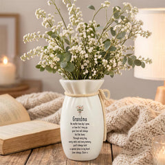 A white vase with a heartfelt message, "Grandma", sits on a wooden table, surrounded by a bouquet of white flowers and a book.