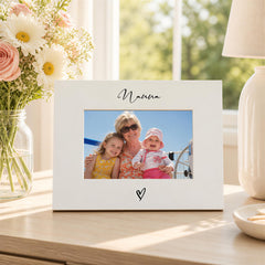 A white picture frame with a photo of a woman and two children, likely a family portrait, sits on a wooden surface. The frame has a heart symbol on the bottom right corner. In the background, there is a vase filled with white flowers and a lamp.