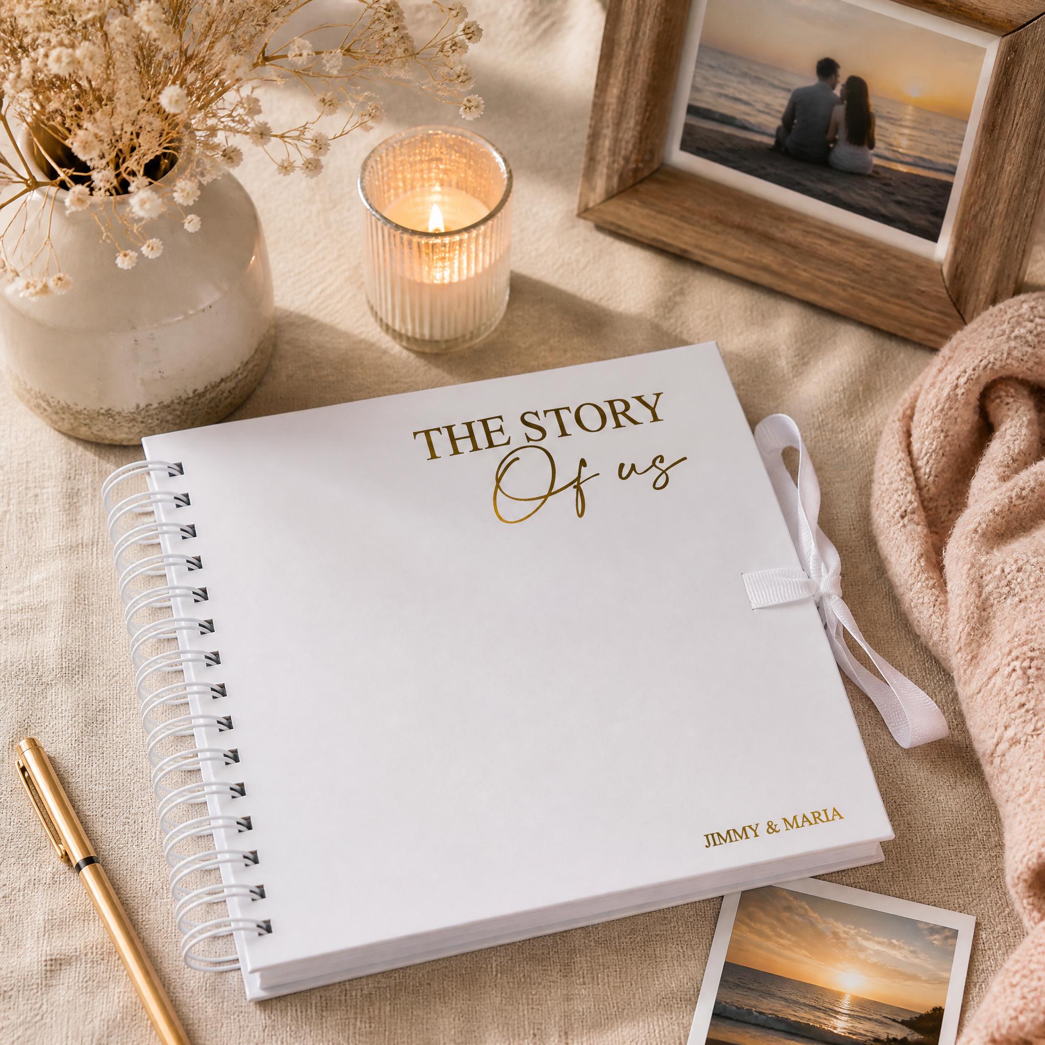 a white spiral-bound notebook with the text "THE STORY OF US" printed on the cover, placed on a beige surface. There is also a framed photograph of a couple, a candle, and a pen nearby.