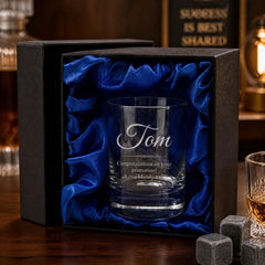 A personalized whiskey glass with the name "Tom" engraved on it, placed in a black box with a blue satin lining. The glass is surrounded by whiskey glasses and a bottle, suggesting a celebration or special occasion.