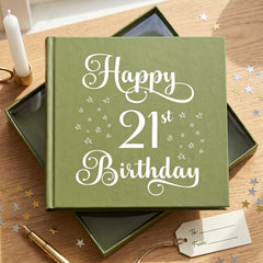 A green book titled "Happy 21st Birthday" with white stars on the cover, placed on a wooden surface with scattered gold stars.