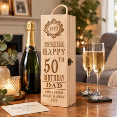 A wooden gift box with a personalized message, a bottle of champagne, and two glasses of champagne on a wooden table.