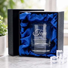 A personalized glass with the name "Tom" engraved on it, placed in a black box with a blue satin lining. The glass is surrounded by ice cubes, and there is a potted plant in the background.