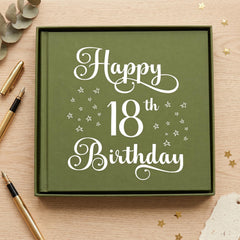 A green book with the text "Happy 18th Birthday" written on it, surrounded by gold pens and scattered gold stars on a wooden surface.