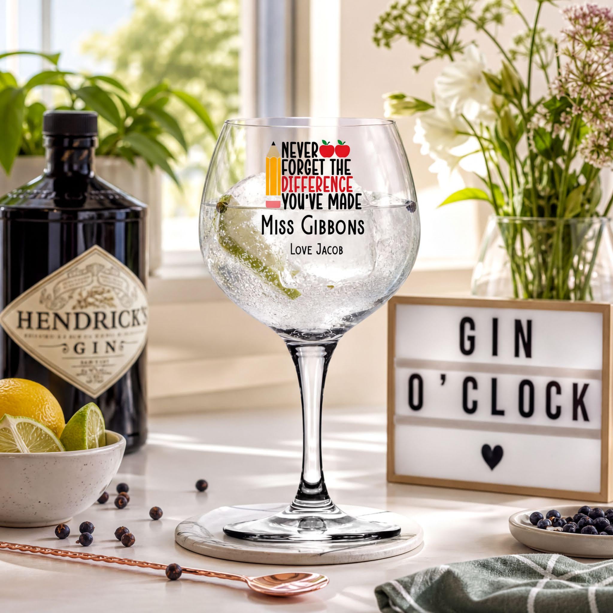 a table with a gin bottle, a gin glass with a message, a bottle of Hendrick's Gin, a bowl of fruit, a bottle of gin, a glass of water, a vase of flowers, a sign, and a spoon.