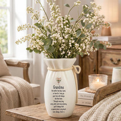 A white vase with a floral arrangement sits on a wooden table, accompanied by a candle and a book.