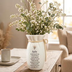 A white vase with a bouquet of white flowers sits on a wooden table, accompanied by a mug and a small plant.