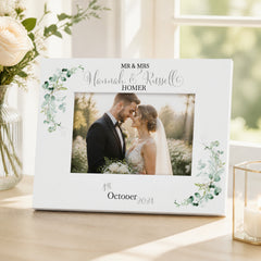 A white picture frame with a photo of a couple, surrounded by green leaves and flowers, sits on a table. The frame has a personalized message that reads "Mr & Mrs Hannah & Russell Homer" and "October 2023".