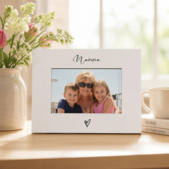 A white picture frame with a photo of a woman and two children, placed on a wooden surface next to a vase of flowers and a mug.