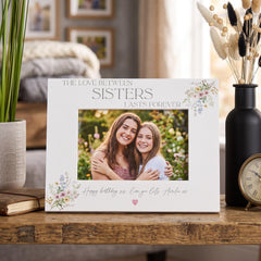 A white picture frame with a photo of two smiling women, placed on a wooden surface. The frame has a floral design and the text "The love between sisters lasts forever" written on it.