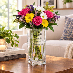 A clear glass vase filled with a colorful bouquet of pink roses, yellow daisies, and purple and white flowers sits on a wooden table. The vase has a personalized message that reads "Mr & Mrs [Name] 18th October 2020".