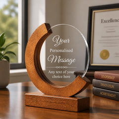 A wooden award or plaque with a clear glass front, featuring a personalized message and a wooden base. The award is placed on a wooden desk, with a stack of books and a plant in the background.