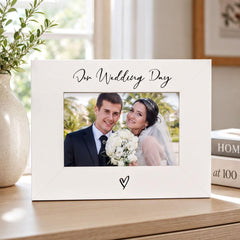 A white picture frame with a photo of a couple, presumably a bride and groom, inside it. The frame has a heart symbol at the bottom. The background of the image is blurred, but it appears to be a room with a window and some books.