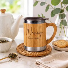 A wooden mug with a metal lid, featuring the name "Nanny" and the phrase "Love Jamie" engraved on it, sits on a woven coaster on a kitchen counter. The mug is accompanied by a teapot, a bowl of tea leaves, and a plate of cookies.