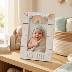 A baby is sitting in a white wooden frame with a rainbow-colored bead decoration, surrounded by baby-related items such as a stuffed animal and a basket.