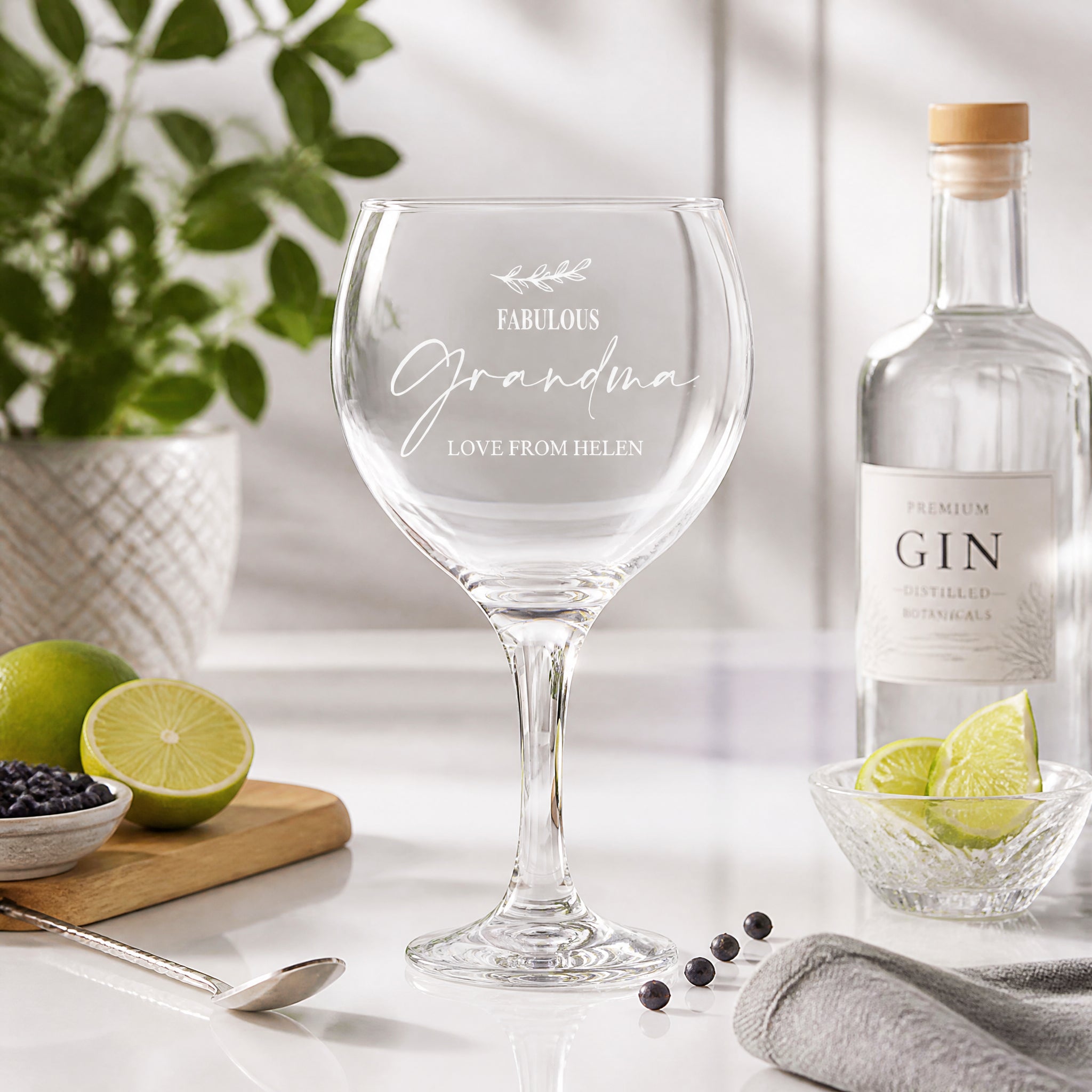 a wine glass with a personalized message, a bottle of gin, and some fruits and a spoon on a table.