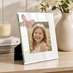 A young girl with a bright smile is the focus of a white picture frame with a butterfly decoration, placed on a wooden surface. In the background, there are books and a vase with flowers, creating a cozy and inviting atmosphere.