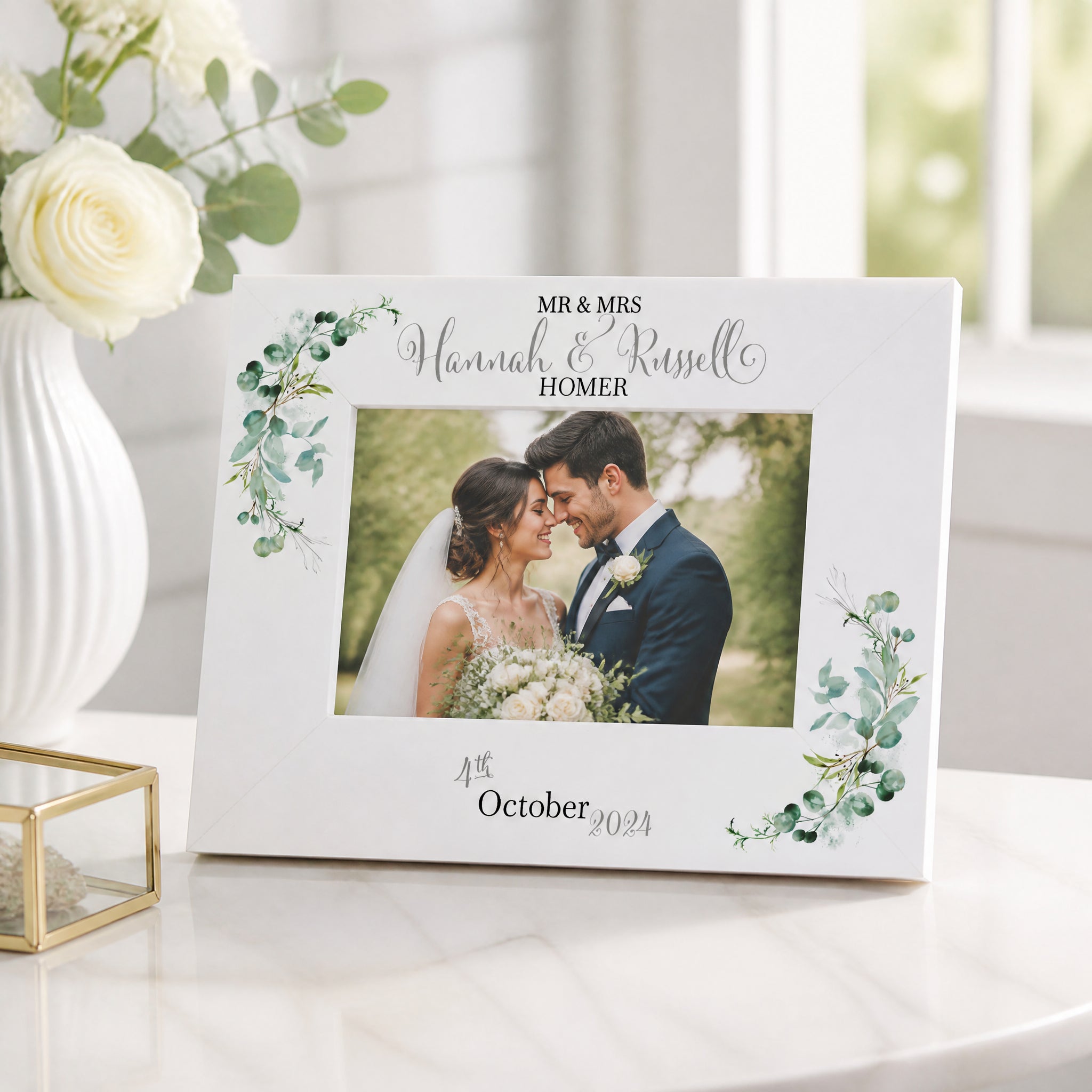 A white picture frame with a photo of a bride and groom, surrounded by green leaves and flowers, sits on a white table. The frame has a decorative border and the names "Mr & Mrs" and "Hannah & Russell" written on it.