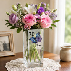 A clear glass vase filled with a bouquet of pink roses, purple roses, and white lilies sits on a lace doily on a wooden table. Next to the vase is a small ceramic jar and a framed photo.