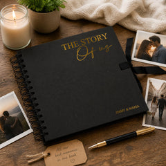 a black spiral-bound notebook with gold lettering, a pen, a candle, and some photographs arranged on a wooden surface.
