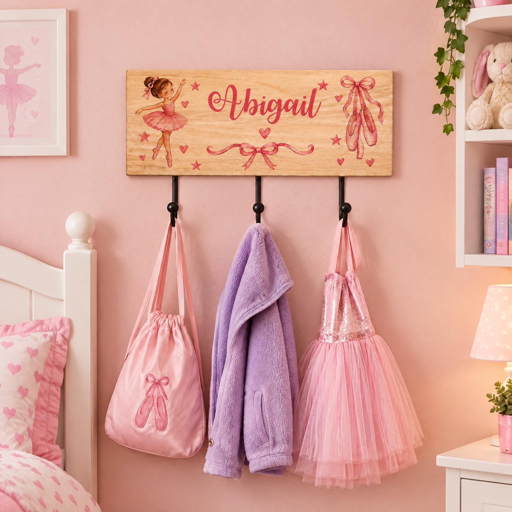 A wooden coat rack with three hangers displaying a pink tutu, a purple jacket, and a pink tulle dress. Above the coat rack is a wooden sign with a ballet girl illustration and the name "Abigail".