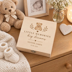 A wooden box with a teddy bear and a heart-shaped ornament on a dresser, along with a photo frame and a vase of white flowers.