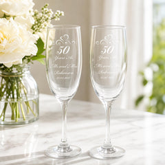 two champagne glasses with the inscription "50 Years As Mr and Mrs Robinson" on them, placed on a marble countertop next to a vase of white flowers.