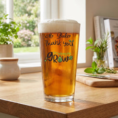 A glass of beer with a personalized message on it, sitting on a wooden table near a window and some plants.
