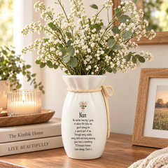 A white vase with a floral arrangement sits on a wooden surface, accompanied by a candle and a book titled "LIVE BEAUTIFULLY".