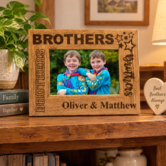 A wooden picture frame with a photo of two smiling children, likely siblings, inside it. The frame has the text "BROTHERS" and "Oliver & Matthew" engraved on it. The frame is placed on a wooden shelf, and there is a heart-shaped wooden object to the right of the frame.