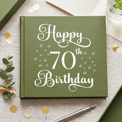 A green book titled "Happy 70th Birthday" with white text and stars on the cover, surrounded by confetti, a pen, and a glass of water on a wooden surface.