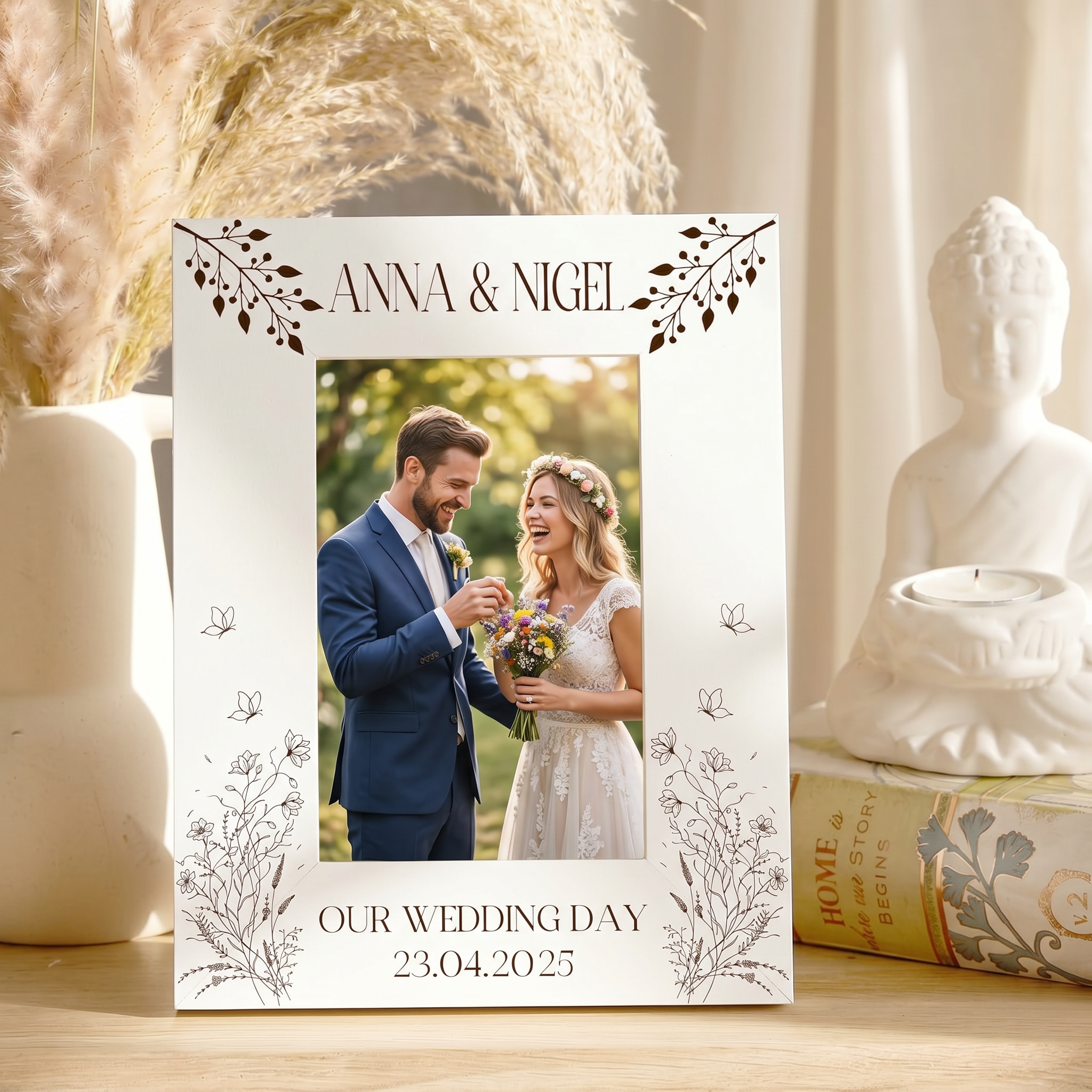 A white wedding picture frame with the names 'Anna & Nigel' and the date '23.04.25' is displayed on a wooden surface, surrounded by decorative items such as a Buddha statue and a vase.