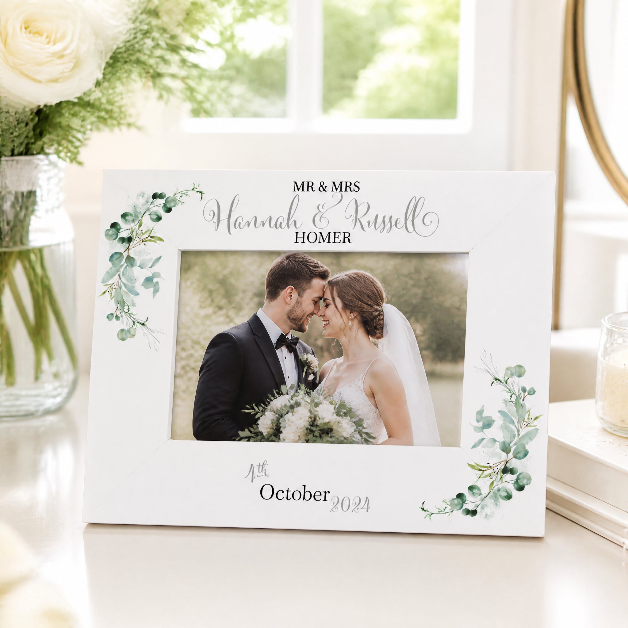 A white picture frame with a photo of a bride and groom, surrounded by green leaves and flowers, sits on a table. The frame has a decorative border and the names "Mr & Mrs Hannah & Russell" written on it.