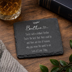 A slate coaster with a heartfelt message about a brother, placed on a wooden surface next to a glass of amber liquid and a book.