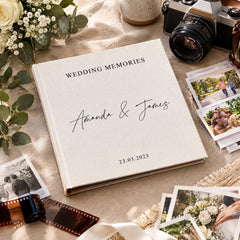a wedding album titled "Wedding Memories" with the names "Amanda & James" written on it, placed on a table surrounded by various wedding-related items such as a camera, film, and photographs.